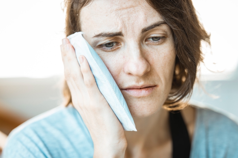 A woman holds a cold pack up to the side of her face, she looks to be in pain